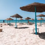 Beach scene with straw umbrellas, white sand, and clear blue sea under a bright summer sky.